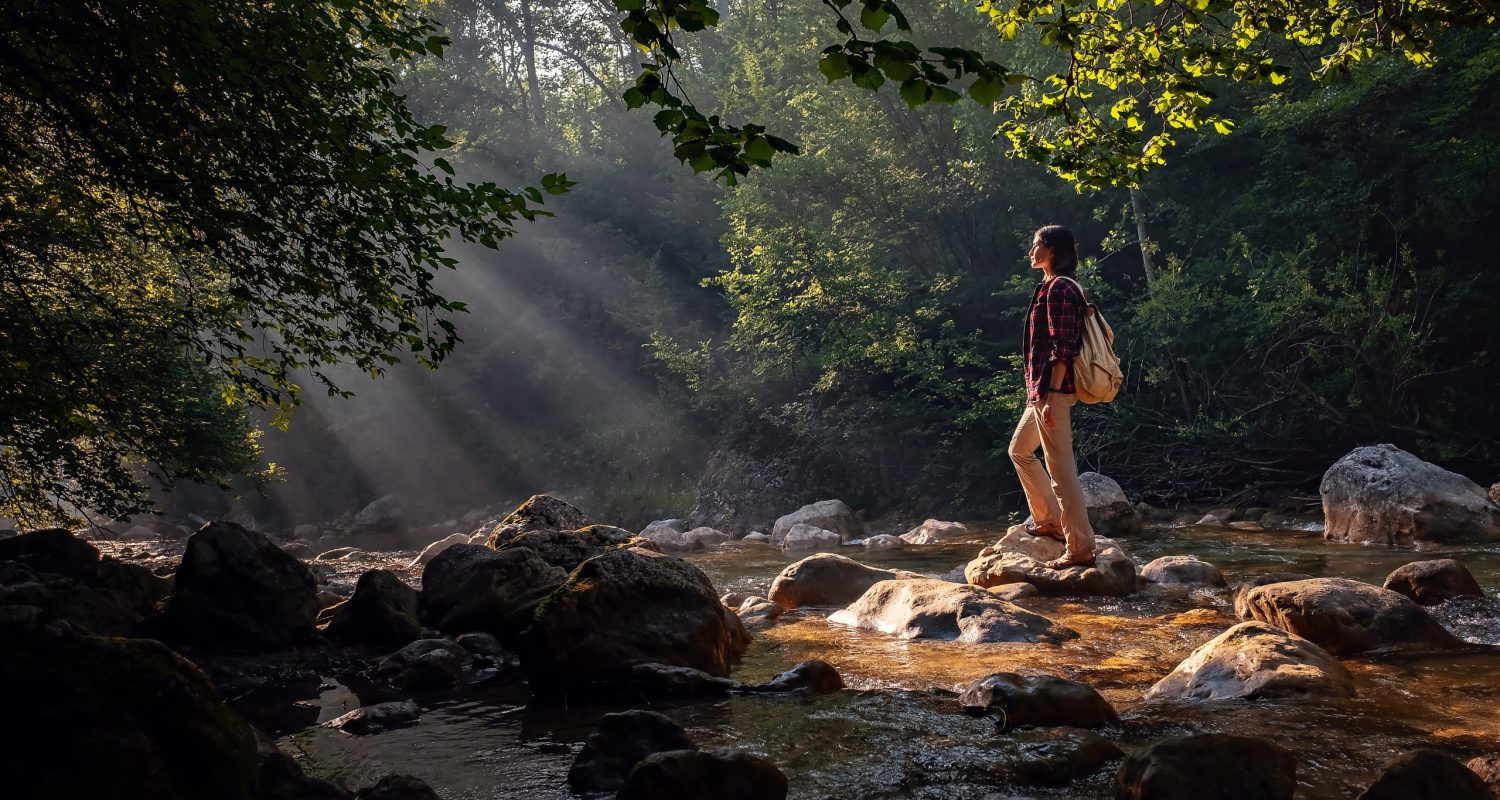 A woman explores new, magical, and fantastic places around the world, surrounded by nature and spreading her arms to breathe and relax. Female hiker crossing the forest creek.